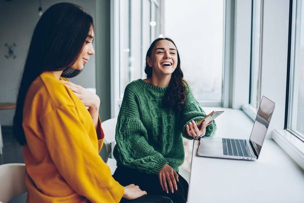 Overjoyed female laughing at funny content from network sitting next to colleague using modern gadgets and wifi, positive women enjoying happy about completing task together sitting at office - Фото, изображение