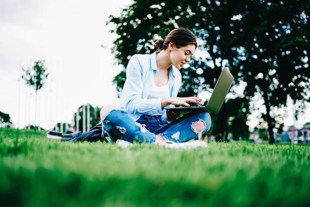 Skilled female freelancer doing remote job via laptop computer in park recreating on spring day, woman studying online courses on typing netbook spending time on green grass in university campus - 写真・画像