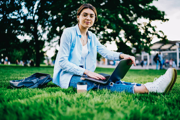 Portrait of young female student in eyewear sitting on grass with laptop computer for online studying, hipster girl looking at camera spending spring free time in park with netbook and wifi connection - Valokuva, kuva
