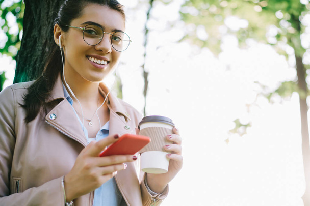 Happy young millennial woman looking at camera satisfied with good sound in earphones and joying music on leisure, portrait of cheerful hipster girl listening playlist songs via mobile phone in park - 写真・画像
