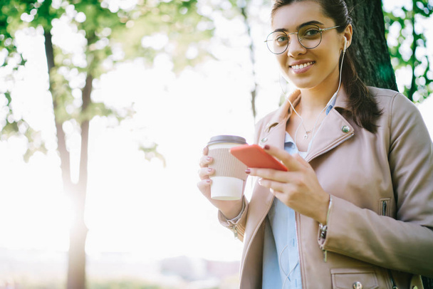 Half length portrait of smiling hipster girl entertaining with playlist songs in earphones having coffee break in park, young millennial woman looking at camera enjoy radio broadcasting via smartphone - Foto, Bild