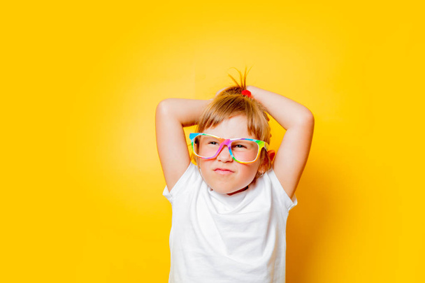 Portrait of emotional toddler girl in white t-shirt and glasses on yellow background. - Фото, изображение