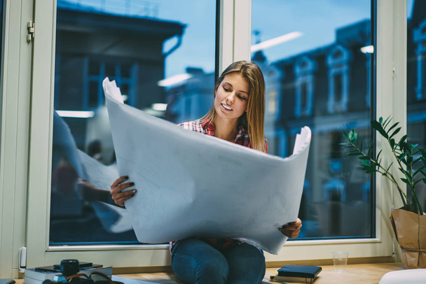 Cheerful young woman holding architectural sketch with graphic sitting on windowsill in modern office during overtime work.Positive successful female designer with blank poster in hands has deadline - Valokuva, kuva