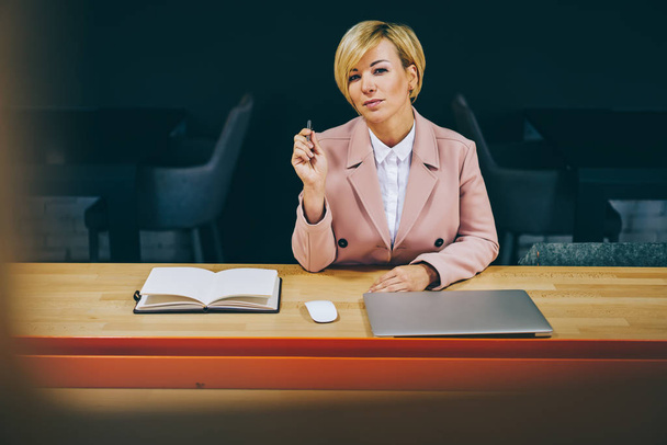 Portrait of experienced female professor 50 years old looking at camera while sitting at wooden table in university.Intelligent mature businesswoman with pen in hands working in office interior - Photo, Image