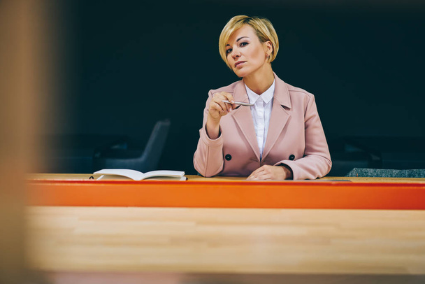Portrait of confident mature businesswoman dressed in elegant wear looking at camera while working in office.Serious experienced female coach sitting at wooden table in college while looking at camera - Photo, image