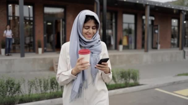 Young woman in gray hijab drinking coffee and looking at smartphone - Metraje, vídeo