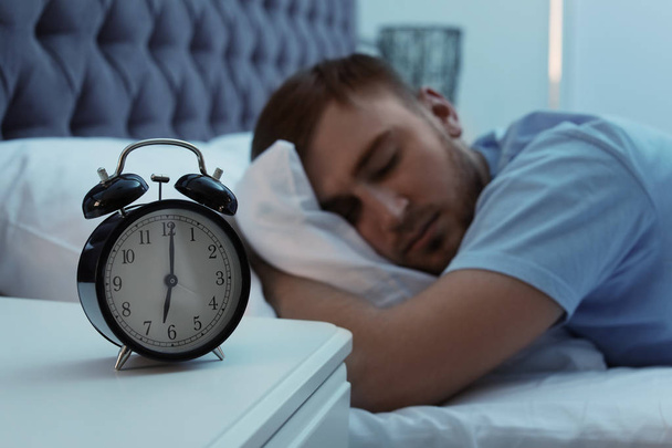 Alarm clock on table and young man sleeping in bed at night - Foto, afbeelding