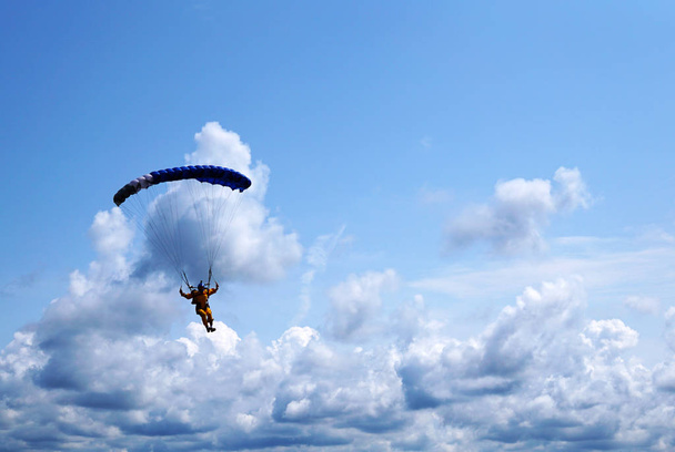 Прыгун с парашютом под тёмно-голубым пологом парашюта на заднем плане голубое небо и облака крупным планом. Силуэт парашютиста с парашютом против неба и грозовыми облаками
.                  - Фото, изображение