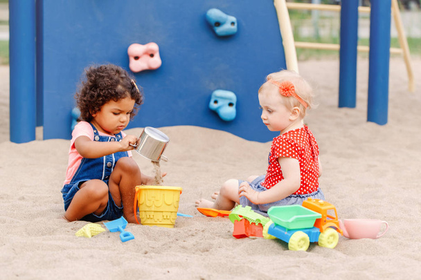 Show me how to do it. Two cute Caucasian and hispanic latin babies children sitting in sandbox playing with plastic colorful toys. Little girls friends having fun together on playground. - Фото, изображение