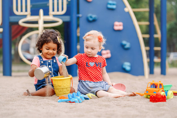 Sandy ground. Two cute Caucasian and hispanic latin babies children sitting in sandbox playing with plastic colorful toys. Little girls friends having fun together on playground. - Фото, изображение