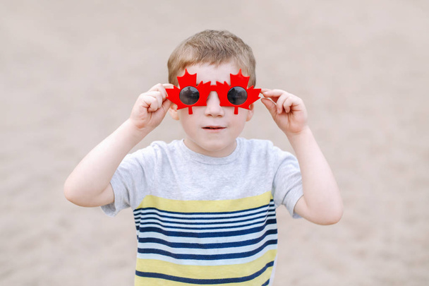 Portrait d'un adorable petit garçon blond caucasien portant des lunettes de soleil canadiennes drôles avec de la feuille d'érable. Enfant célébrant la fête du Canada et s'amusant
.  - Photo, image