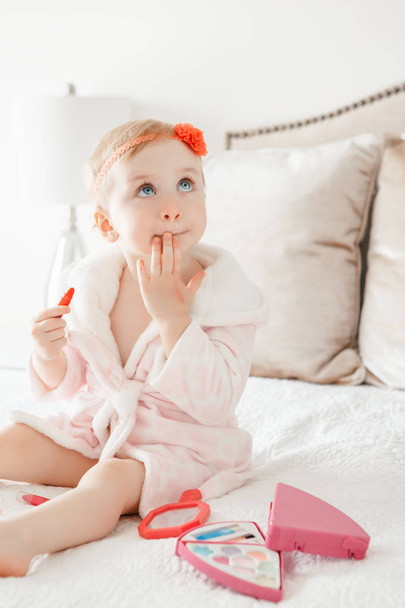 Cute asorable funny Caucasian blonde baby girl 2 years old in bathrobe doing applying makeup, looking into mirror and sitting on her bed in bedroom at home. Happy childhood concept. - Фото, зображення
