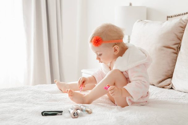 Schattig leuk blond Kaukasische babymeisje schilderij nagels zittend op bed thuis. Vrouwelijke kind doen manicure. Peuter spelen met behulp van cosmetica.  - Foto, afbeelding