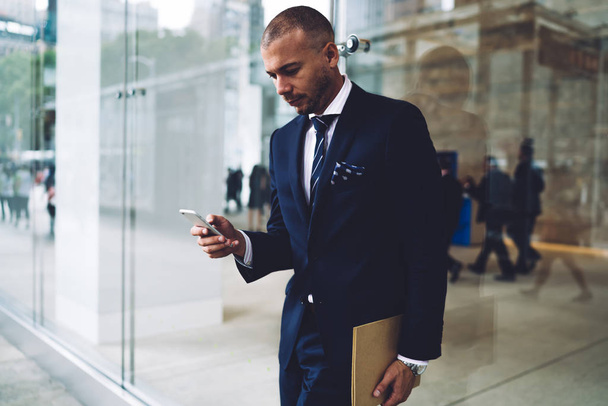 Confident pensive proud ceo reading financial news on website on smartphone device while standing near office building.Serious businessman sending sms message on telephone using 4G internet - Foto, Imagen