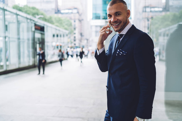 Cheerful young entrepreneur dressed in formal wear smiling at camera while communicating on smartphone standing on street.Positive successful businessman calling on mobile and talking in roaming - Photo, Image