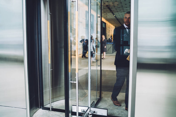 Experienced owner of financial company dressed in formal wear going out on street from modern office building on coffee break.Young businessman standing on entrance and opening glass door - Photo, Image