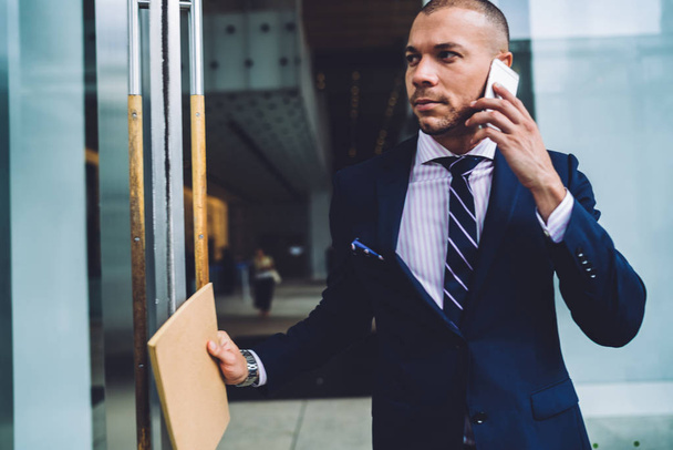 Serious businessman in formal wear going out from office building and communicating during phone conversation on smartphone.Confident young entrepreneur in suit talking on cellular - Photo, image