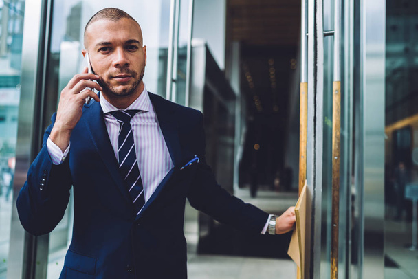 Portrait of confident businessman opening door and go out of office building while calling on smartphone device.Young entrepreneur dressed in formal wear communicating on mobile phone - 写真・画像