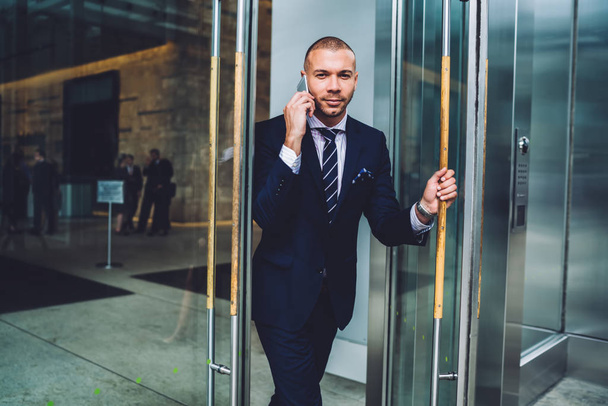正式な摩耗で成功した起業家の肖像画は、スマート フォンで電話での会話中にオフィス会社と開口部のガラス扉から行きます。ビジネスマンの休憩に行く携帯電話で呼び出す - 写真・画像