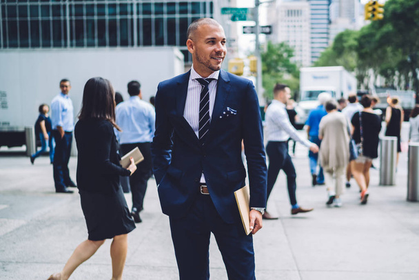 Cheerful young businessman dressed in formal wear holding folder in hand and smiling away while standing outdoors on street of modern city.Positive entrepreneur in suit in downtown - Fotografie, Obrázek