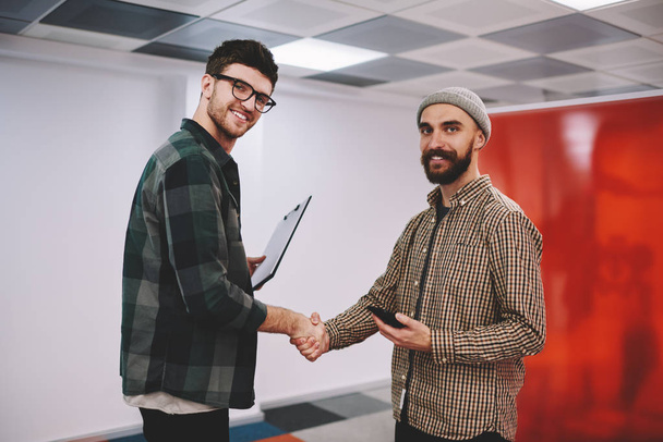 Portrait of successful young men dressed in casual wear smiling at camera while handshaking after business deal.Prosperous hipster designers have concluded an agreement standing in modern office - Foto, imagen