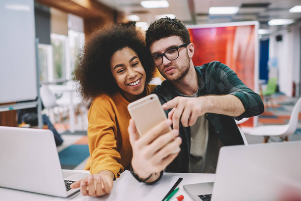 Positive african american hipster girl together with cheerful caucasian young man dressed in casual wear smiling at front camera of modern smartphone and making selfie for publication in internet blog - Foto, Imagem