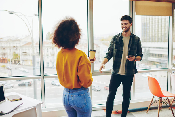 Two cheerful multicultural colleagues dressed in casual wear having fun during break in office.African american hipster girl communicating with positive caucasian young man enjoying leisure time - Photo, image