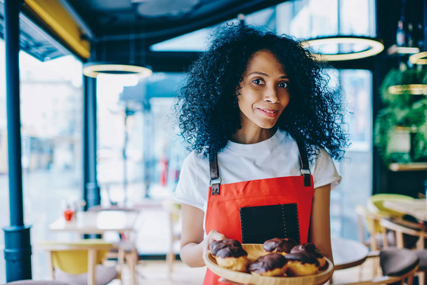 Half length portrait of successful female pastry cook in apron holding plate with tasty chocolate cakes standing in own bakery shop.Beautiful dark skinned confectioner showing biscuit desserts - Фото, зображення
