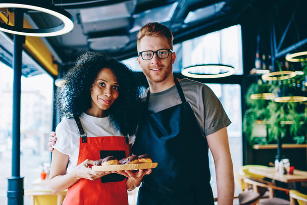 Portrait of multicultural positive employees of bakery shop dressed in aprons showing plate with sweet cakes.Partnership of smiling male barista and professional pastry chef with chocolate desserts - Zdjęcie, obraz
