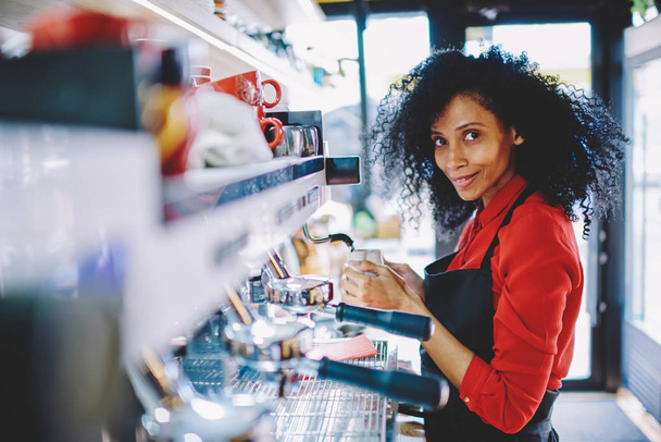 Portrait of successful african american young woman dressed in uniform working barista in own cafeteria and preparing tasty coffee beverage for client on modern machine developing own business - Foto, Bild