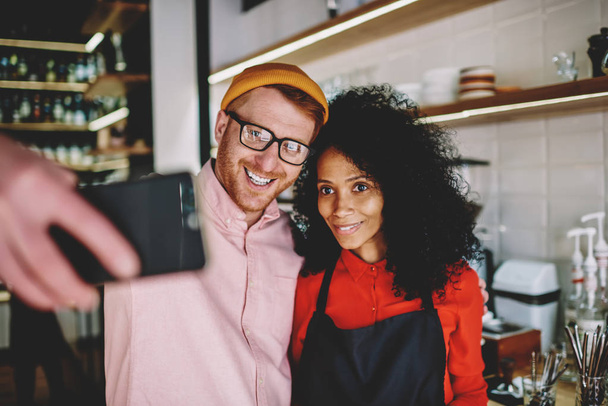 Cheerful african american professional barista making selfie photo together with caucasian friend on smartphone.Positive dark skinned waitress taking photo on front camera of celluar with hipster guy - Фото, изображение
