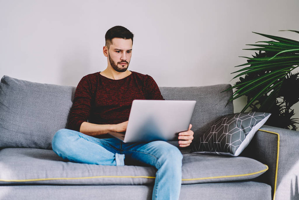 Pensive bearded young man freelancer watching tutorial online on internet websites using modern netbook with high speed internet connection resting on comfortable couch in modern apartment interior - Foto, imagen