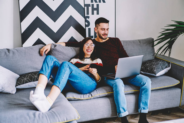 Positive couple in love enjoying free time on cozy sofa and watching funny movie on modern laptop using wireless internet connection in apartment.Young wife and husband resting at home - Photo, image