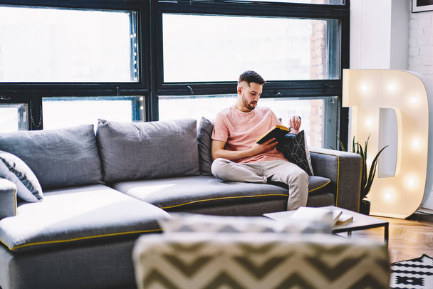 Pensive young man resting at home on comfortable sofa and reading book in modern apartment with stylish interior.Hipster guy enjoying literature plot sitting on cozy couch in design flat - Zdjęcie, obraz