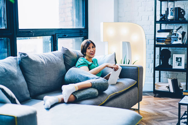 Portrait of happy young woman lying on comfortable sofa and spending leisure time on reading book in apartment.Positive beautiful hipster girl with bestseller smiling at camera relaxing at home - Photo, Image