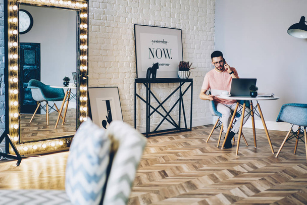 Young man talking on smartphone while working remotely at modern netbook with wireless internet in modern apartment with stylish design interior.Male freelancer calling on telephone at home - Valokuva, kuva