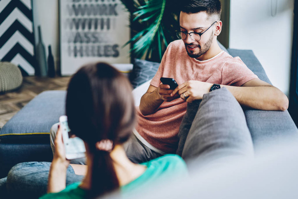 Cheerful young man relaxing together with girlfriend on comfortable couch and discussing blog with news in social networks while messaging in online chat on modern smartphones using wireless internet - Fotó, kép