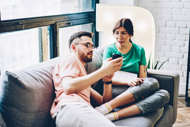 Young man showing internet publication on website on modern smartphone to girlfriend relaxing on comfortable couch in common stylish apartment.Husband with wife watching videos in blog on cellular - Foto, immagini