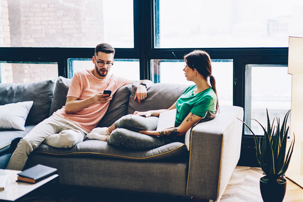 Young marriage spending time together in modern apartment with stylish interior.Young man searching website on smartphone device using home internet while sitting on sofa with girlfriend in flat - Foto, Imagen