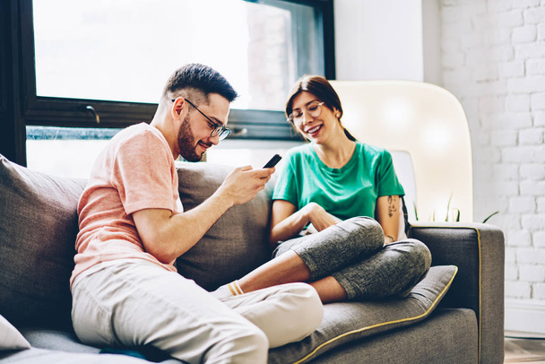 Positive couple in love laughing and discussing funny story in blog on smartphone using home internet in stylish apartment.Cheerful wife with husband relaxing on couch and spending time together - Foto, Imagem