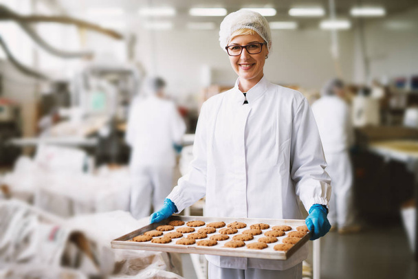 młody zadowolony pracownik kobieta trzyma świeżo upieczone ciasteczka na blachę w zakładzie produkcji żywności - Zdjęcie, obraz