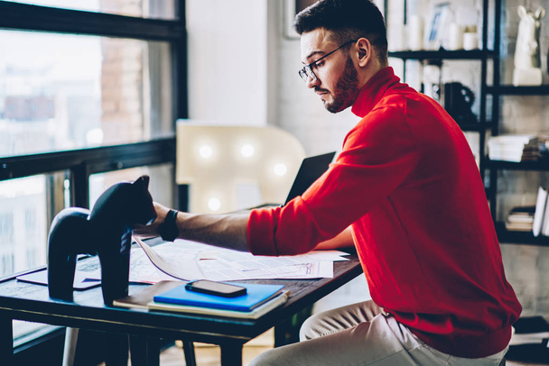 Concentrated young man in eyeglasses working on developing design project sitting at desktop in modern studio.Pensive creative hipster guy planning organizational process in modern apartment - Фото, изображение