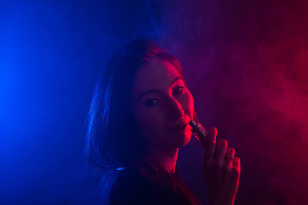 Portrait of young woman in neon red and blue smoke with vape or e-cigarettes - Foto, imagen