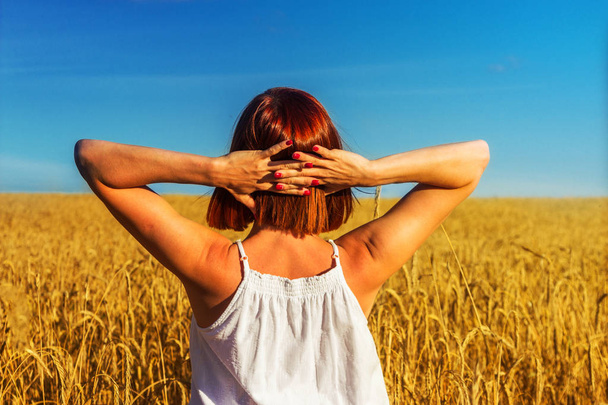 Freedom. Red-haired tanned girl stands in the middle of a wheat field at sunset - Photo, Image