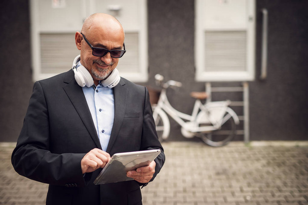 Улыбающийся взрослый бизнесмен пользуется планшетом с наушниками на шее
 - Фото, изображение