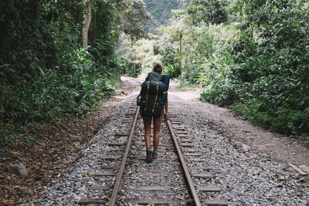 Back view of professional female hiker with tourist backpack exploring wilderness environment during hiking trekking on railway in destination of mountains and green vegetation to discover new places - 写真・画像
