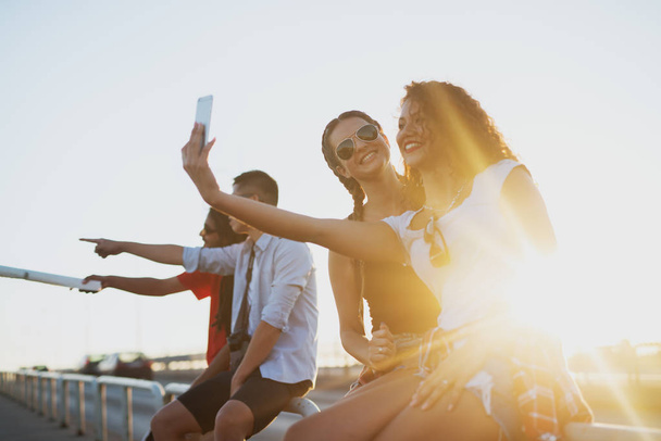 Gruppe junger, glücklicher Freunde, die in der Stadt unterwegs sind, auf der Brüstung einer Brücke sitzen und ein Selfie machen - Foto, Bild