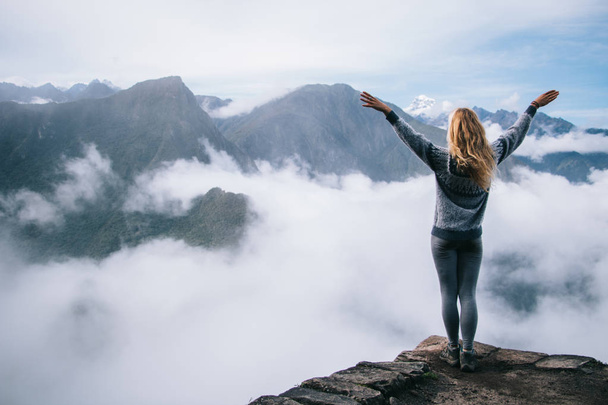 Back view of young woman tourist holding hands up enjoying freedom and travel lifestyle.Female hiker admiring beautiful scenery of green high mountains covered white mist standing on edge of summit - Фото, изображение