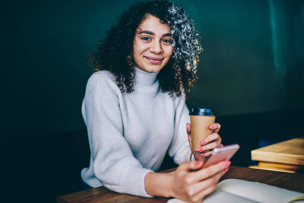 Prosperous millennial female blogger enjoying morning takeaway coffee during checking notification on cellphone, smiling curly woman looking at camera after booking hotel via application at smartphone - Фото, изображение