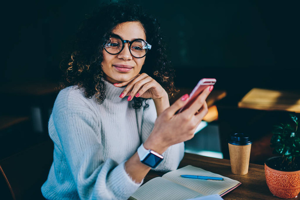 Happy thoughtful woman choosing application for chatting on smartphone sitting with textbook for planning at coffee shop, positive trendy hipster girl in eyewear holding mobile phone and looking away - 写真・画像